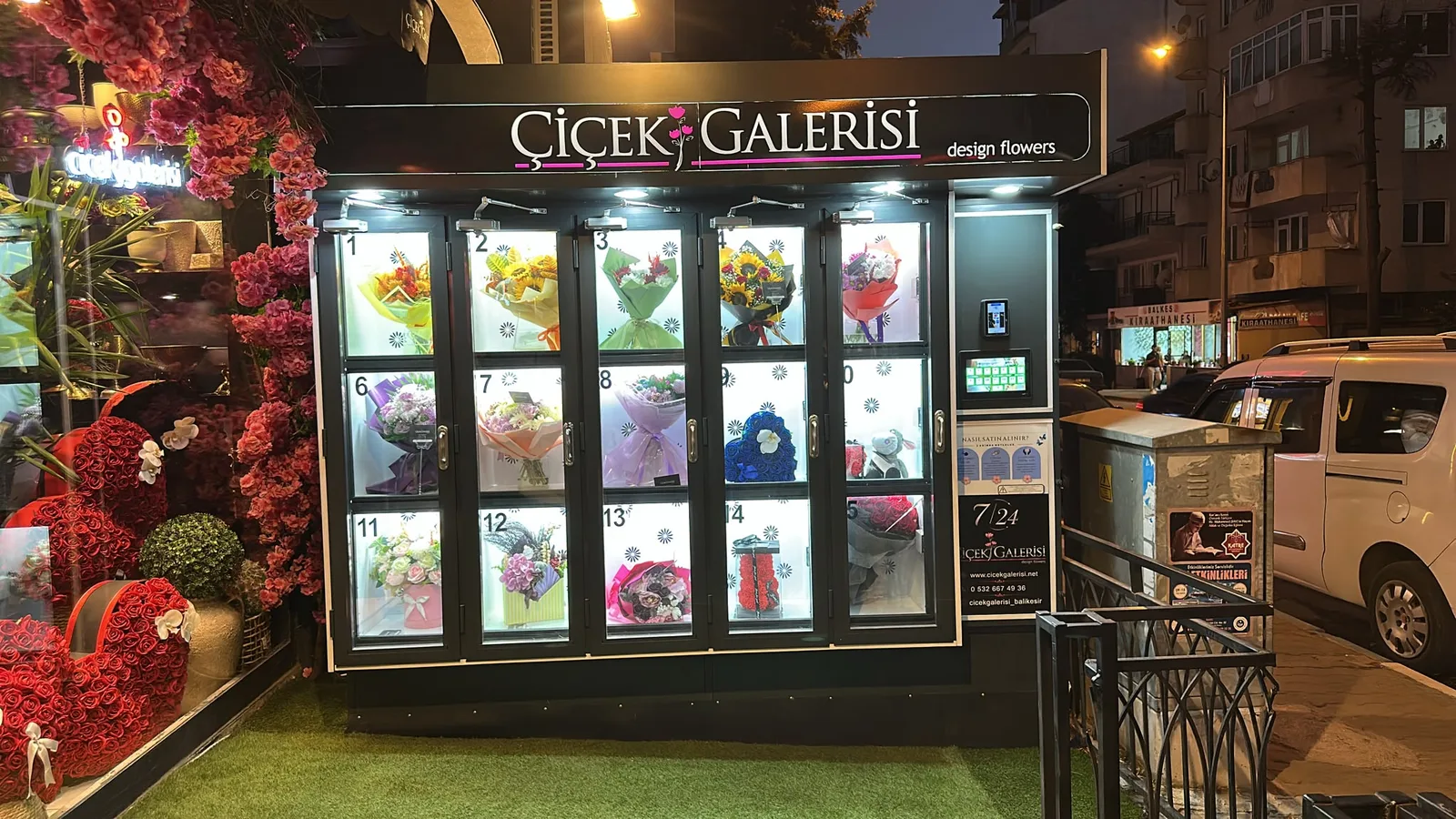 Mehmetçik Caddesi'nde gece: solda dükkânın çiçekli vitrini, sağda otomat ve kaldırım